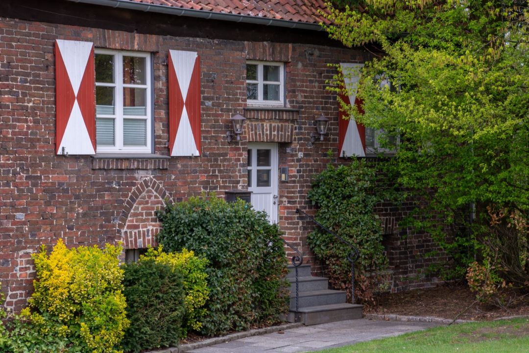 Backsteinhaus mit weiß und rot gestrichenen Fensterläden, davor gewachsene Bepflanzung
